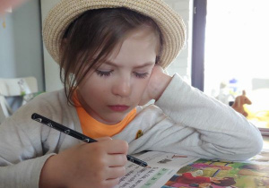 dziewczynka w słomkowym kapeluszu czyta patrząc w książkę i trzymając czarny ołówek